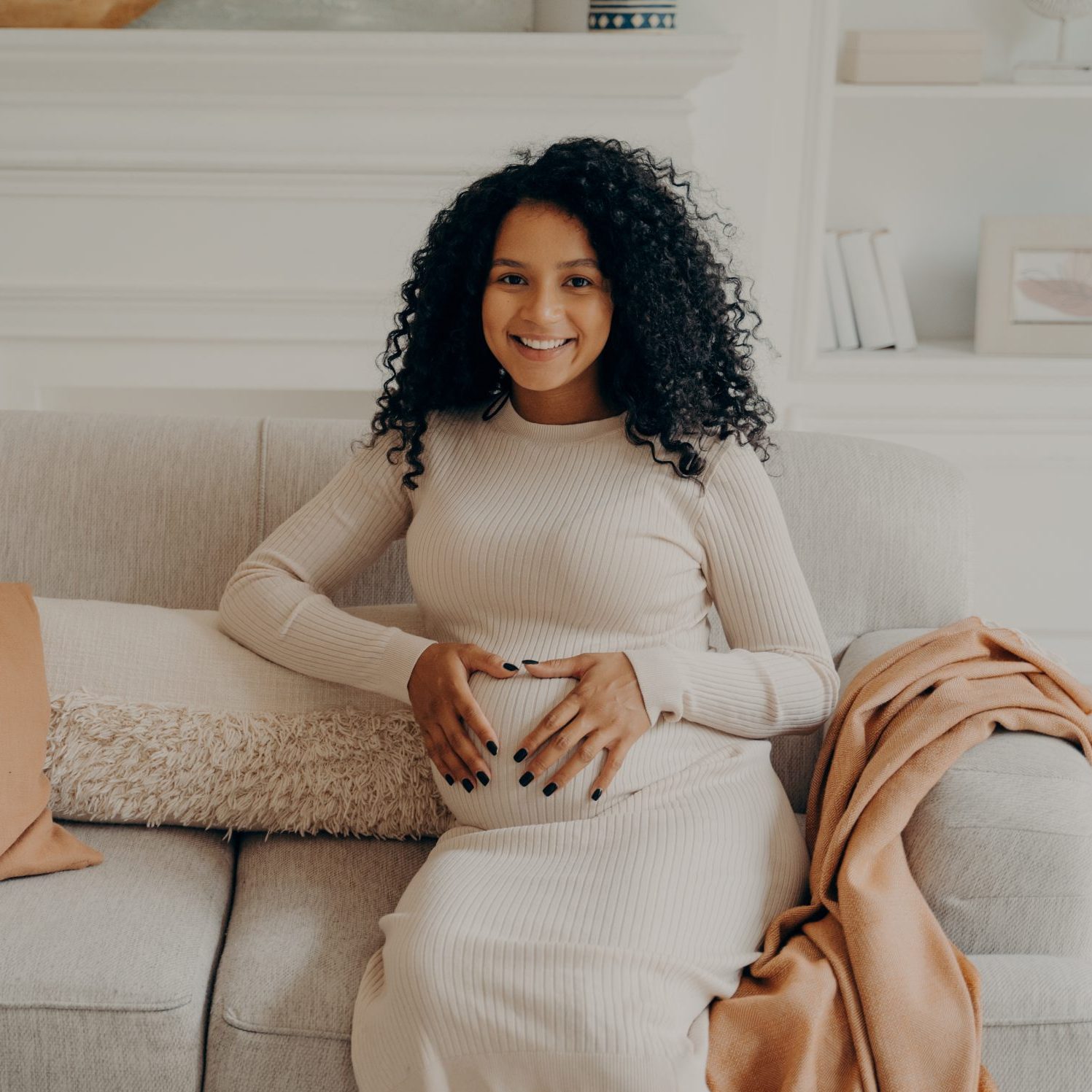 A Black pregnant woman sits on a couch with her hands on her belly